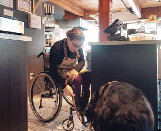 A worker in a boutique leans over in her wheelchair to interact with her service dog. She is wearing glasses and an apron.