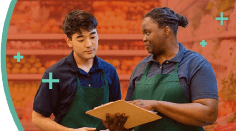 Two grocery store workers wearing aprons review information on a clipboard in the produce section.