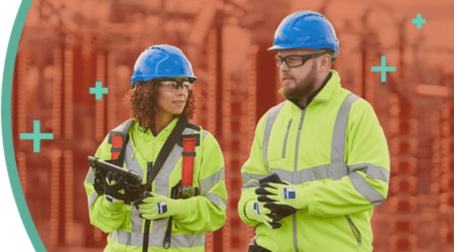 Two construction workers wearing blue hard hats and high-visibility jackets walking and talking, both are holding mobile devices.